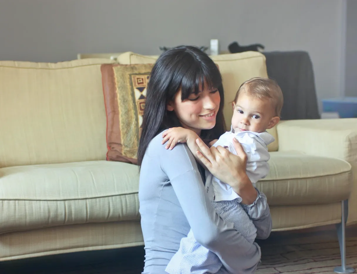 A young au pair lifting a baby in her arms in front of a cream-coloured sofa