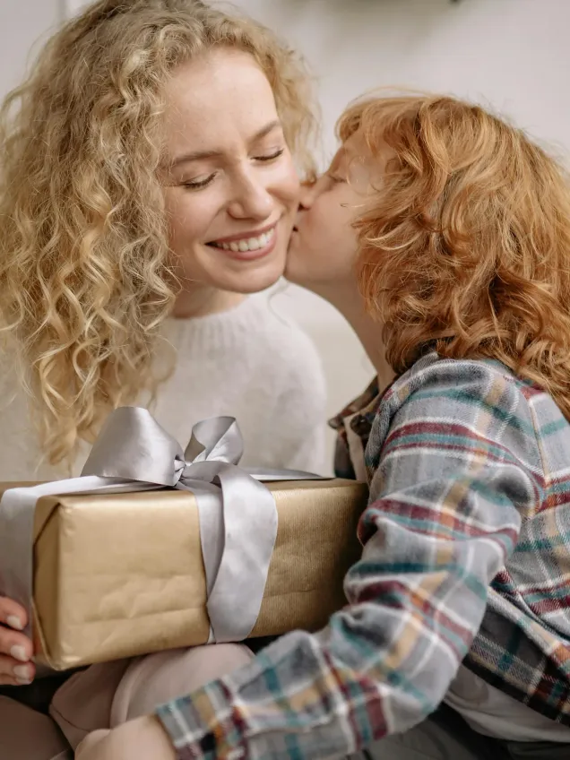 A smiling woman with blonde curls receiving a kiss on the cheek from a child holding a wrapped gift