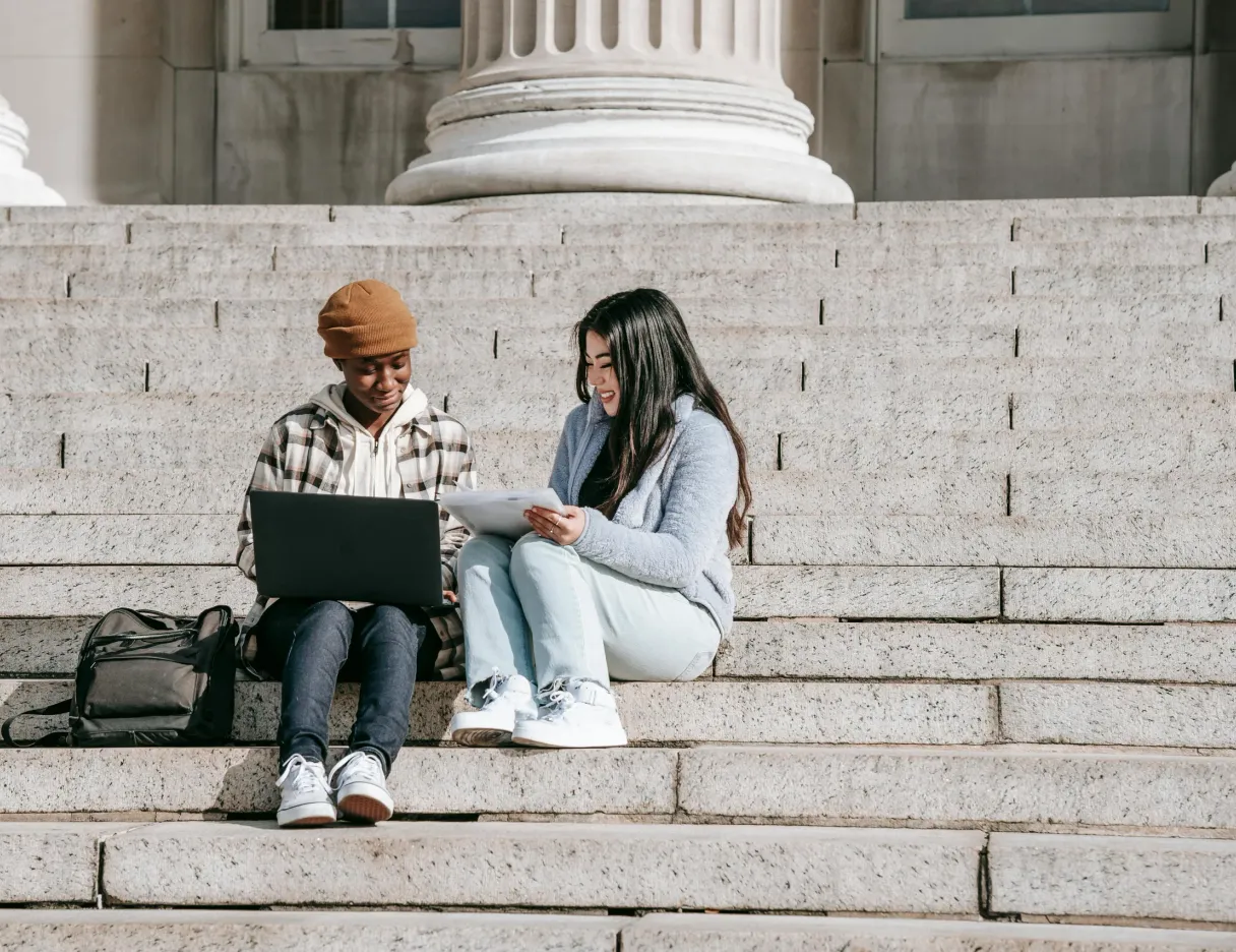 Two students sitting on wide stone steps, looking at a laptop together